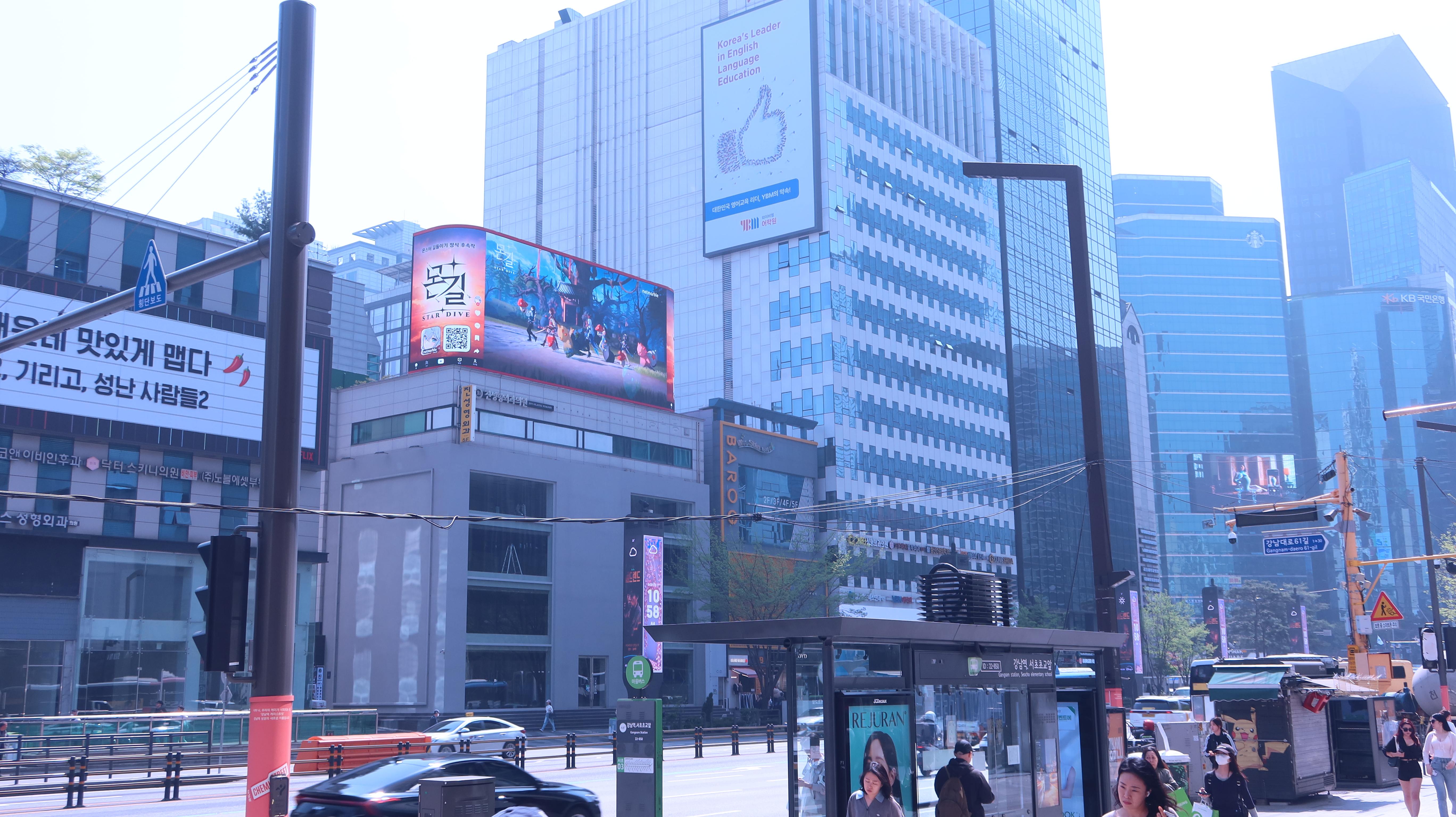 MONGIL: STAR DIVE gameplay creative on the curved rooftop screen near Gangnam Station, with office towers and a bus stop below, Seoul