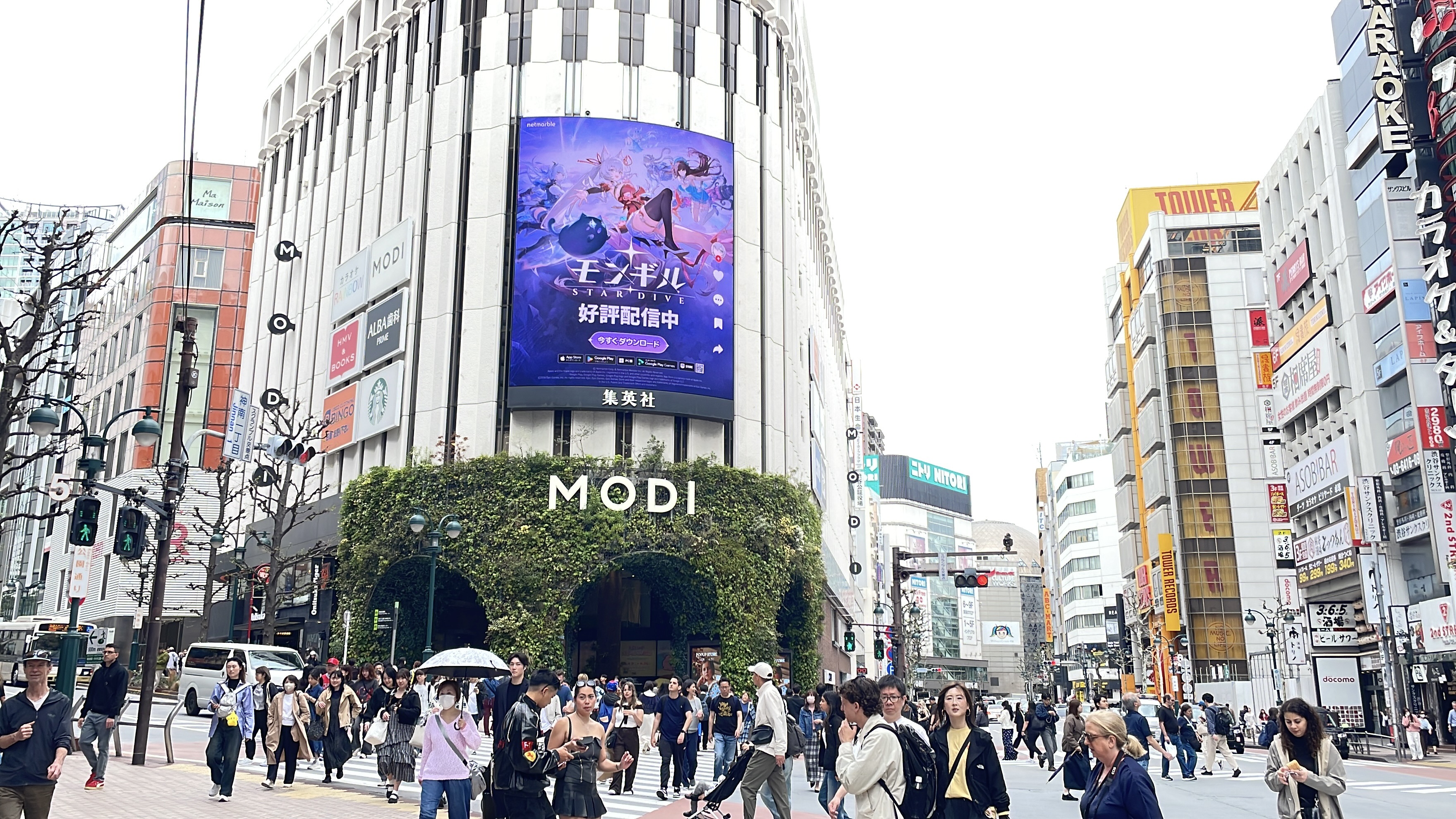 Japanese MONGIL: STAR DIVE key art on the curved MODI screen as crowds cross the street, Shibuya, Tokyo