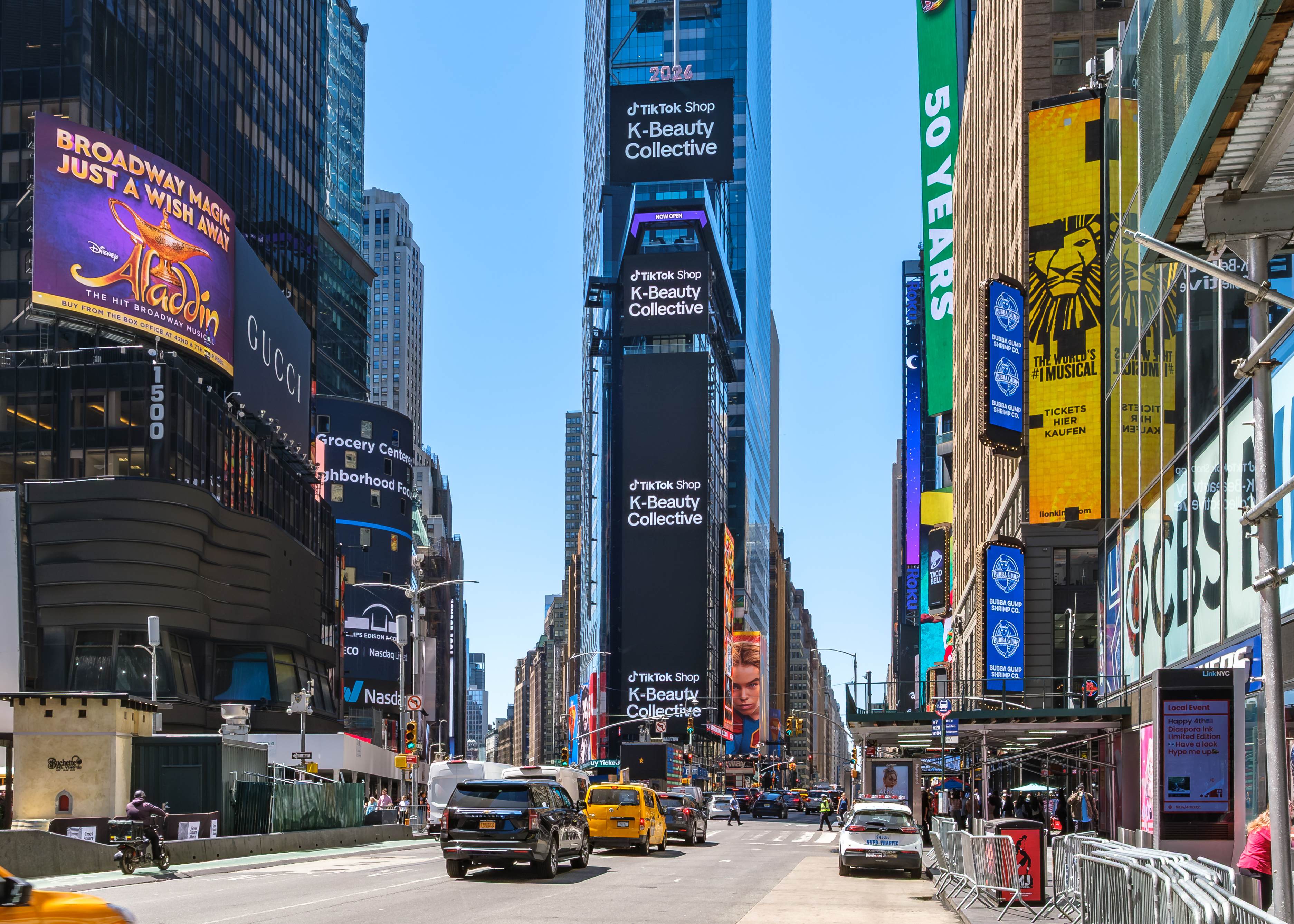 Traffic and pedestrians on Seventh Avenue beneath the TikTok Shop K-Beauty Collective vertical screens and surrounding Times Square media
